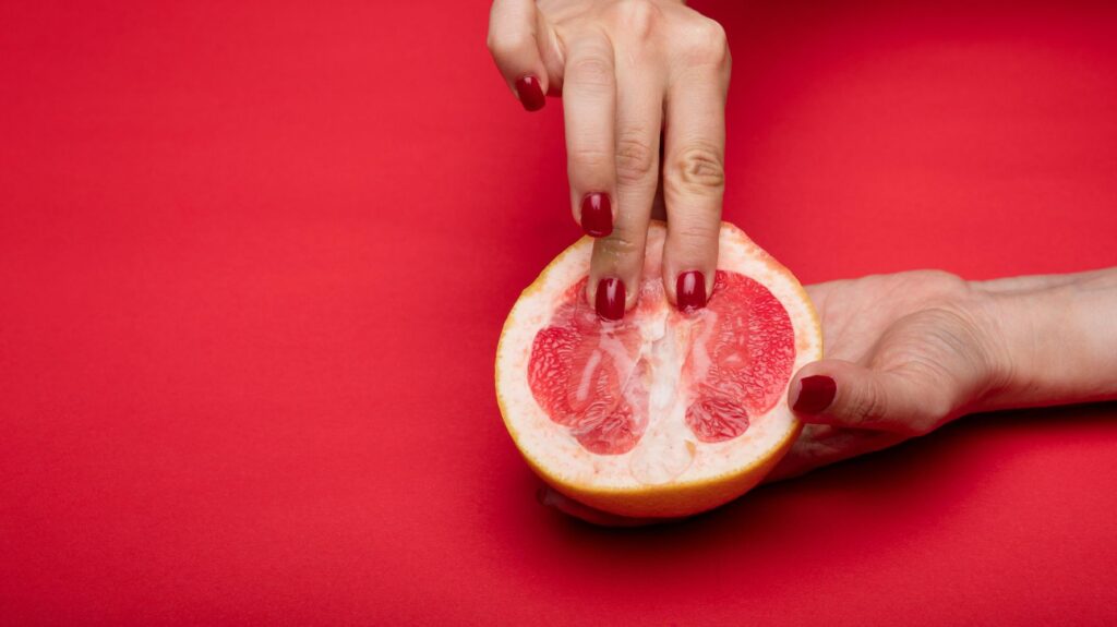 mãos de mulher com unhas vermelha acariciando uma toranja que remete ao clitóris