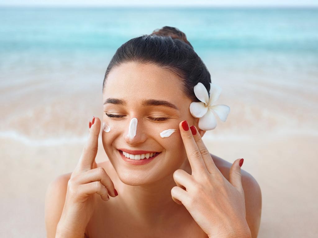 mulher com cabelo preso em coque e uma flor na orelha aplicando protetor solar nas bochechas e nariz