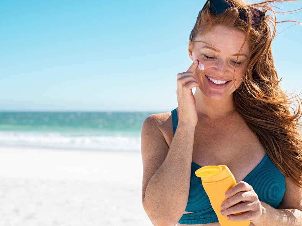 mulher ruiva na praia sorrindo com um frasco de protetor solar na mão