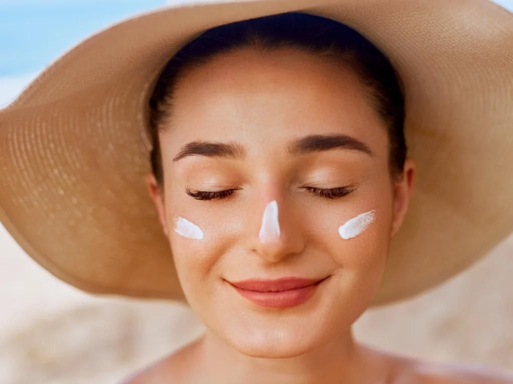 mulher com protetor solar no rosto de lhos fechados e chapéu