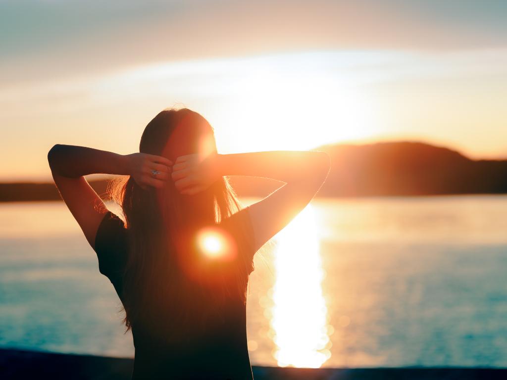 mulher de costas com as mãos na cabeça olhando para o horizonte na praia