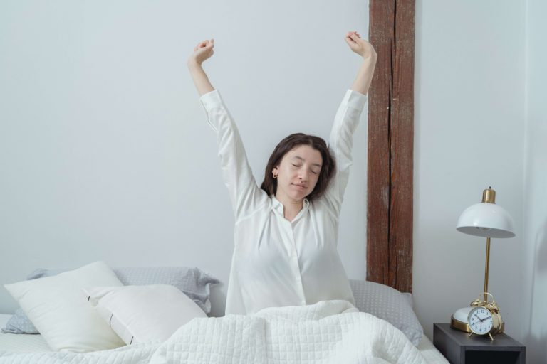 Mulher sentada na cama, embaixo do edredom, com os braços para cima, se alongando.