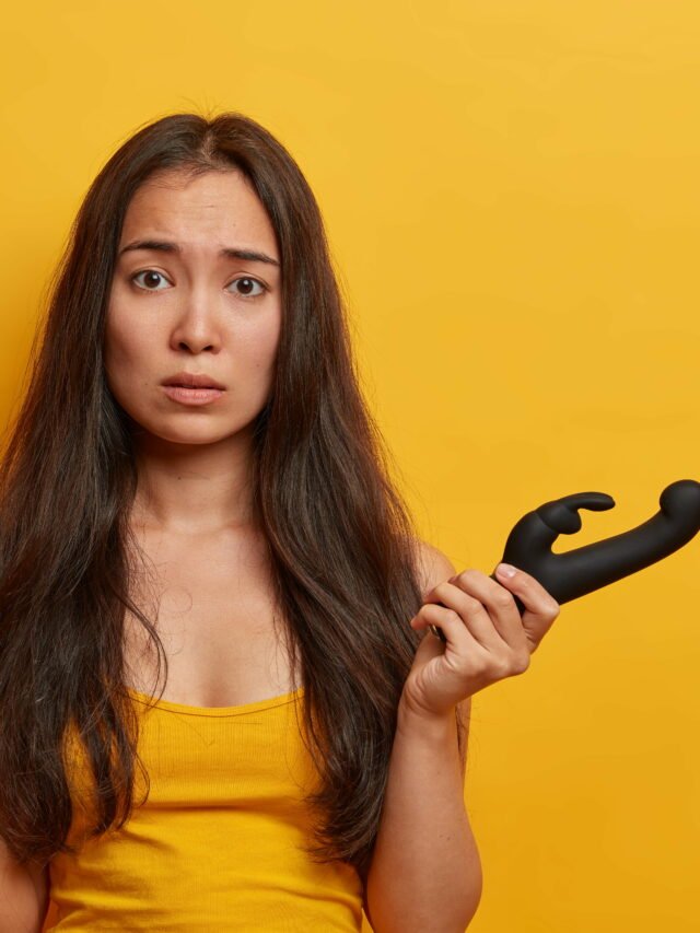 Sex toys, masturbation devices. Worried Asian woman holds silicone dildo and butt plug, prepares for intimate sex games, pleasures herself with vibrator, stands against yellow background. Intimacy