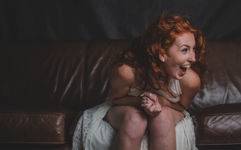 mulher ruiva sentada em um sofá. Ela é ruiva, tem sardinhas no rosto, está com um vestido branco e sorrindo bastante.