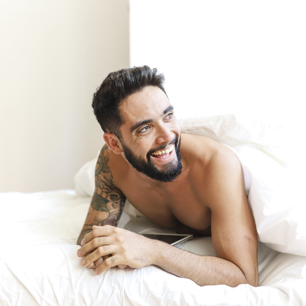 Homem de cabelo e barba preta deitado de bruços, sem camisa, sorrindo, em uma cama com coberta branca cobrindo suas costas.