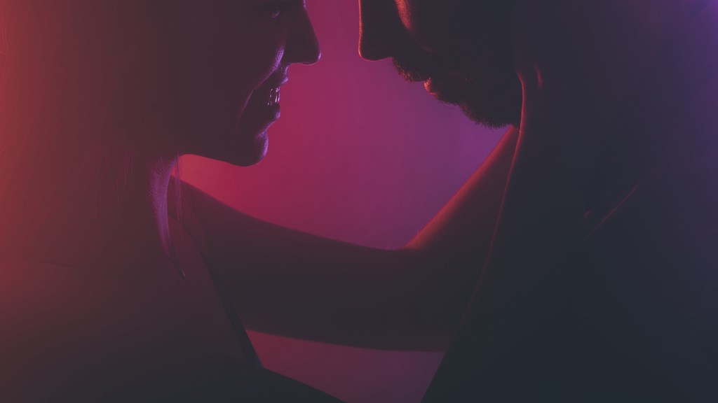 Homem e mulher com rostos bem próximos, em ambiente com baixa iluminação em tom mais próximos do roxo e do rosa, troando olhares sensuais.