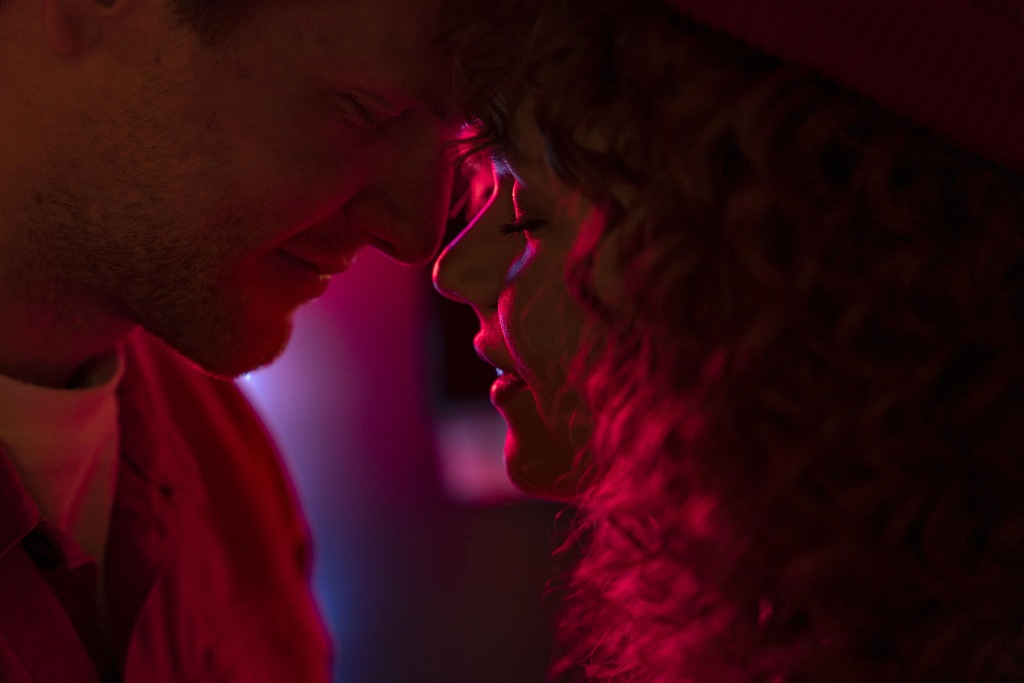 Homem e mulher se encarando, de forma sensual e com os rostos próximos, em ambiente de luz vermelha.