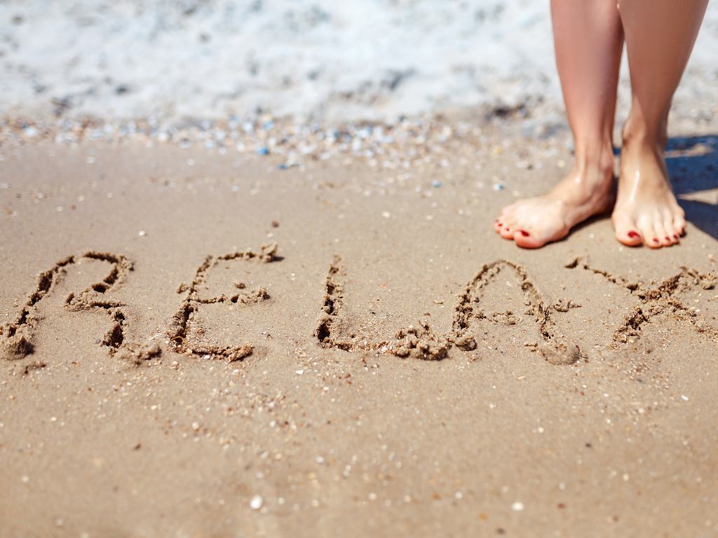 pés de uma mulher na praia com a palavra "relax" escrita na areia