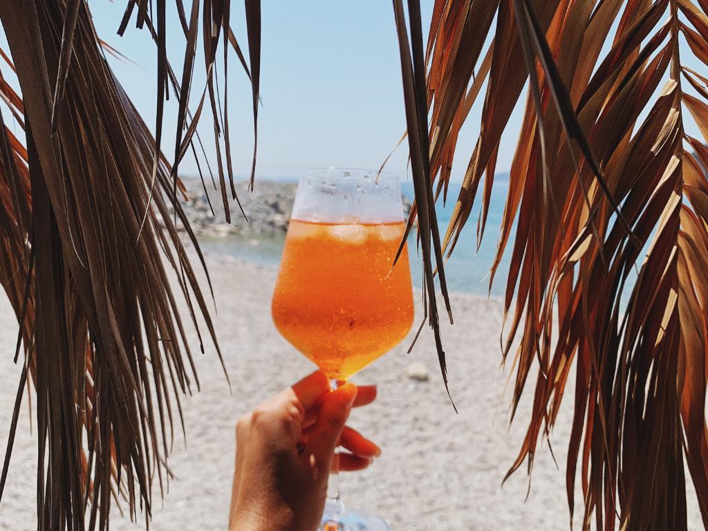 mão segurando um drink laranja na praia com folhas secas dos dois lados