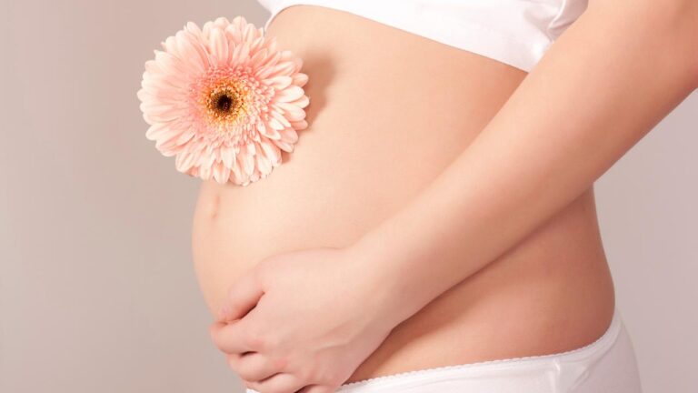 Mulher grávida em pé, com top e calcinha brancos e uma flor em cima da barriga.