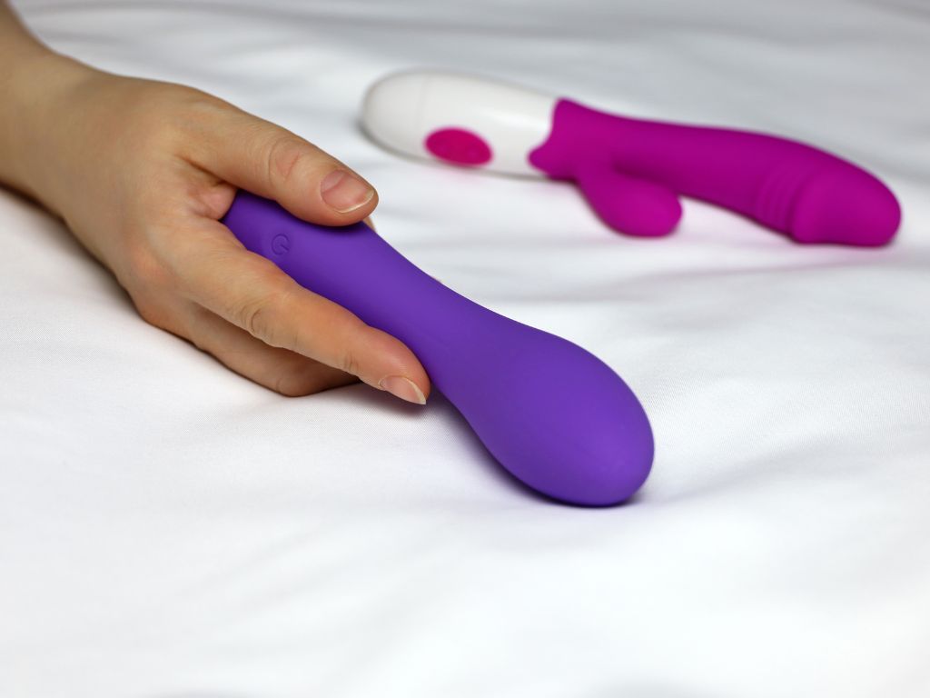 vibradores roxo e rosa na cama com uma mão segurando para ilustrar um post sobre quanto custa um vibrador