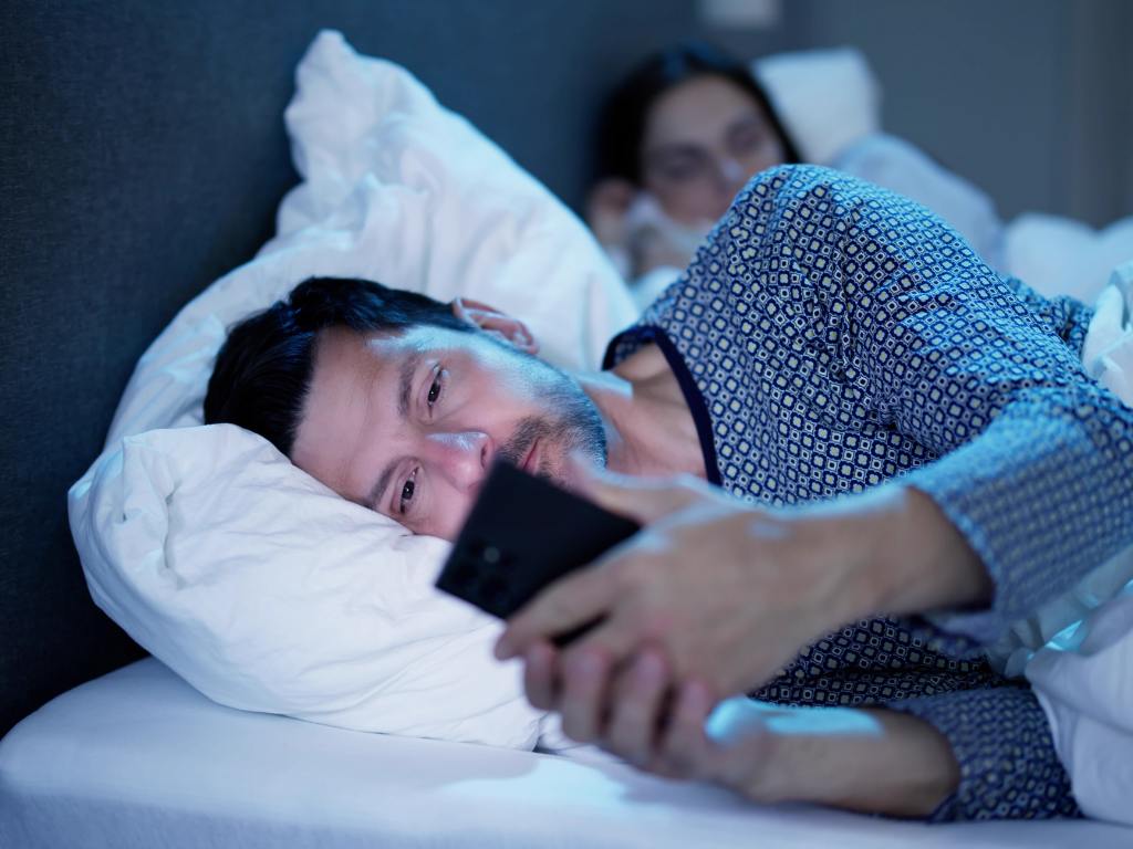 homem com celular na mão na cama praticando traição virtual