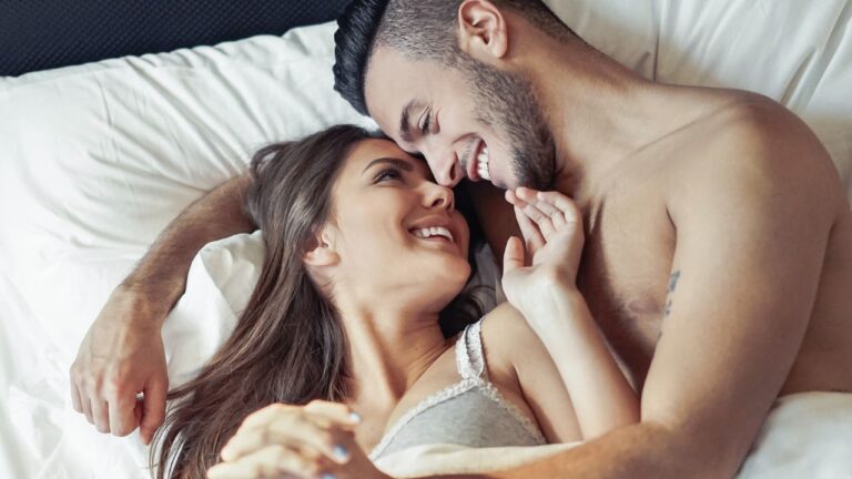 casal na cama sorrindo mulher na frente homem atrás testando posição para engravidar