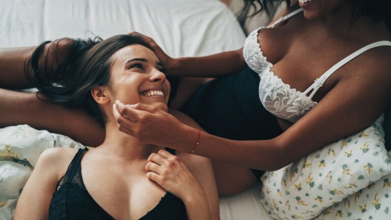 mulher deitada no colo de outro sorrindo na cama, as duas de lingerie