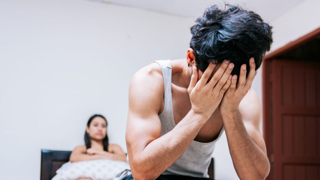 homem em primeiro plano sentada na cama triste e mulher atrás